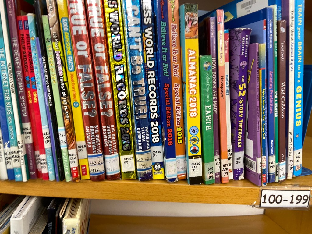 nonfiction library books on a shelf, labels are varied, some printed, some handwritten, some unreadable